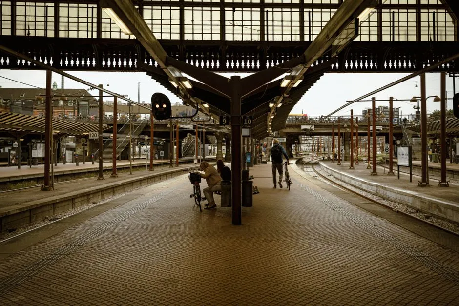 A covered train station platform with an ornate glass canopy overhead and tracks curving into the distance, two people far down the platform each standing beside a bicycle, seen from a distance with no faces visible and no readable signage or logos