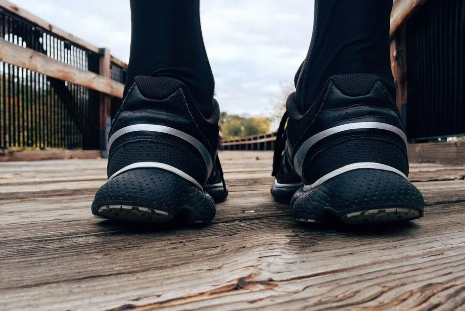 Close-up of a runner's shoes and ankles seen from behind on a wooden boardwalk, framed by a railing leading toward trees in the distance