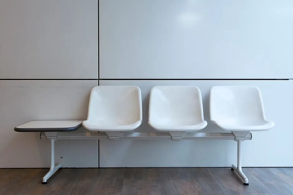 Empty white bench-style waiting room seating with three attached chairs against a plain wall, no people or readable signage visible