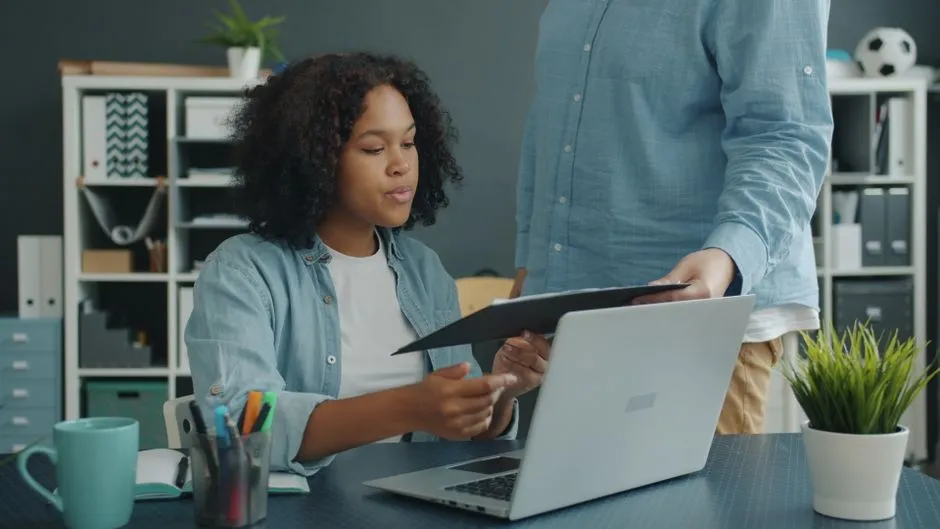 A colleague hands over employment documents at a desk with a laptop in an office