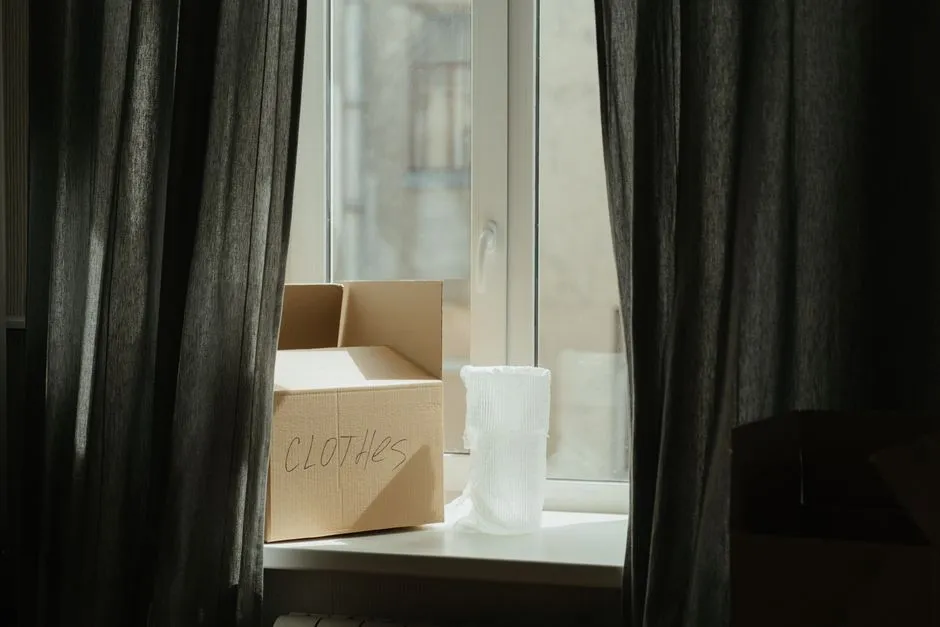 A cardboard moving box labeled clothes on a windowsill beside a window, with heavy curtains partly drawn