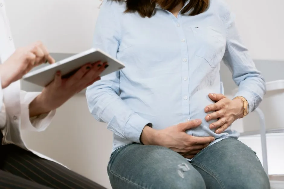 A pregnant woman's torso, hands resting on her belly, while a healthcare provider holds a clipboard nearby, no faces visible