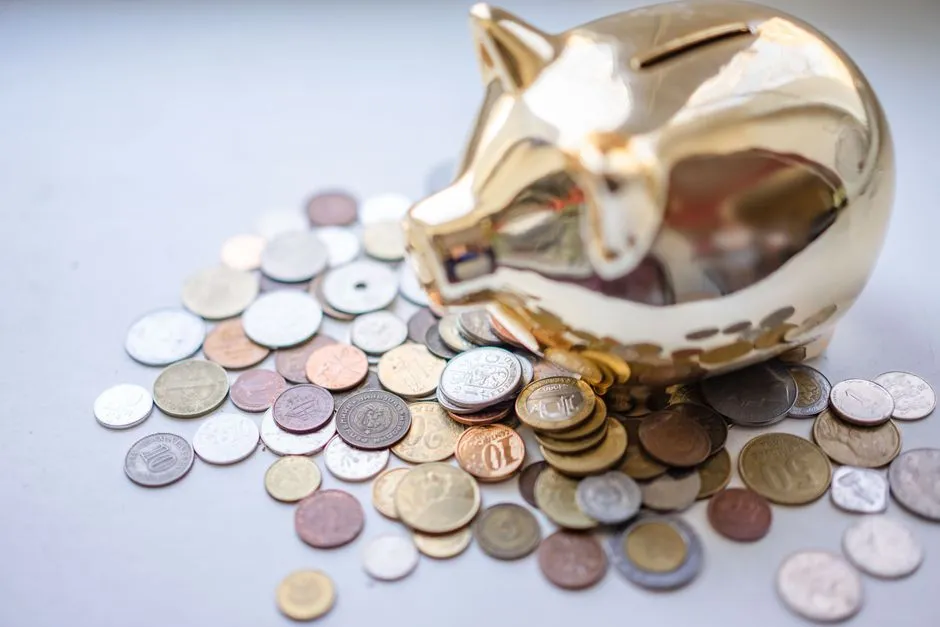 A gold piggy bank surrounded by a scattered pile of coins of different sizes and currencies on a pale surface