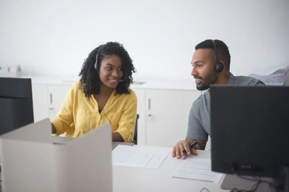 Two office counselors wearing headsets discuss paperwork together at a desk