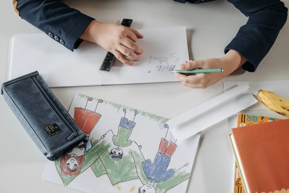 A child's hands drawing at a desk covered in colored pencils, a pencil case, notebooks, and a hand-drawn picture