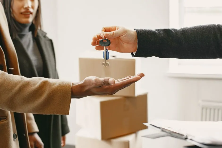 A hand holding out a set of apartment keys toward another person's open palm, with moving boxes in the background